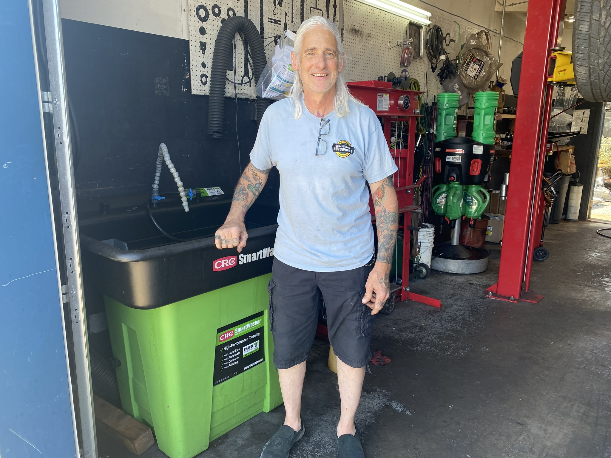 A person with long white hair wearing a blue t-shirt stands in an auto body shop in front of a safe degreaser and a wall of tools and shop parts